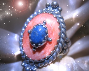 May include: A silver ring with a large, oval, coral-colored stone as the centerpiece. A smaller, blue stone is set in the center of the coral stone. The ring has a detailed, twisted silver band and decorative silver accents.