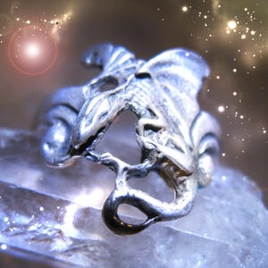 Puede incluir: Un anillo plateado con un diseño de dragón. El anillo presenta un dragón detallado con alas y cola, elaborado en una estética de fantasía estilizada. El anillo se muestra sobre una superficie cristalina transparente.