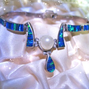 May include: A silver-toned bracelet featuring a series of rectangular blue and green iridescent opal inlays. The center of the bracelet showcases a round white pearl flanked by two angled opal segments, with a single teardrop-shaped opal pendant.
