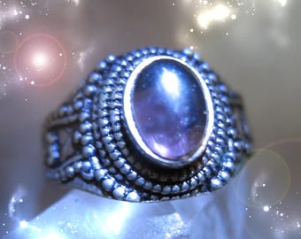 May include: A silver-toned ring with an oval, purple gemstone. The ring has a detailed, textured design around the gemstone and along the band. The ring is set against a soft, blurred background with sparkling highlights.