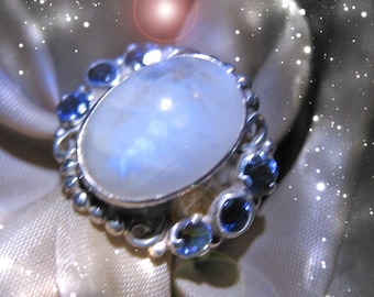 May include: A silver ring featuring a large, oval, iridescent moonstone as the centerpiece. The ring is accented with smaller, round, blue gemstones and intricate silver detailing. The ring is set against a soft, shimmering background.