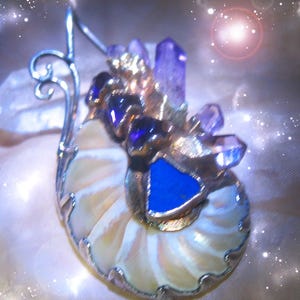 Könnte beinhalten: Handgefertigter Anhänger mit Nautilus-Muschel-Design, verziert mit Amethystkristallen, blauen Edelsteinen und einer silberfarbenen Metalleinfassung. Der Anhänger hat ein einzigartiges, künstlerisches Aussehen, mit Fokus auf natürliche Elemente und einem Hauch von Glanz.
