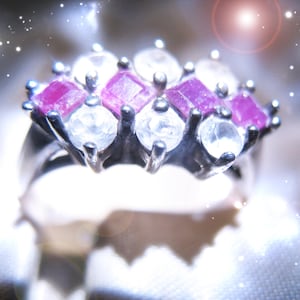 May include: A silver ring with a cluster of gemstones. The ring features a combination of round, clear stones and square, pink stones. The gemstones are set in a silver band, creating a sparkling and elegant design. The ring is a piece of jewelry.
