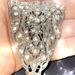 May include: A silver-toned, ornate, vintage brooch with a geometric design. The brooch is covered in clear rhinestones.