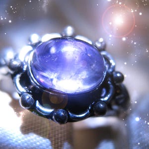 May include: A silver ring featuring a large, oval-shaped, translucent purple cabochon gemstone set in a decorative metal bezel. The setting is adorned with small, rounded metal spheres along the outer edge, creating a textured, metallic frame around the central purple stone.