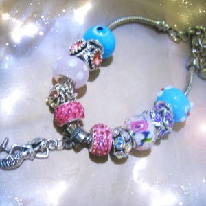 May include: A silver charm bracelet with various colorful beads. The beads include pink, blue, and white designs, with a mermaid charm at the clasp. The bracelet has a shiny, reflective appearance.