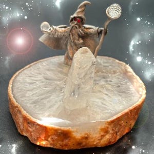 May include: A silver wizard figurine with red eyes and a staff, standing on a white and brown agate slice. The wizard is holding a crystal ball. A large, clear crystal is in front of the wizard.