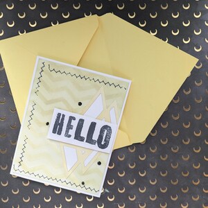 May include: A yellow card with a white border and a geometric design. The card has the word "HELLO" printed in black letters. The card is sitting on a black background with a gold crescent moon pattern.