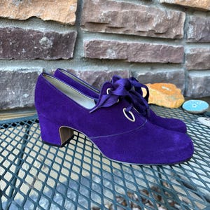 May include: A pair of purple suede shoes with a low heel and lace-up detailing. The shoes have a rounded toe and a small, stacked heel. The laces are a matching purple colour. The shoes are sitting on a metal surface.