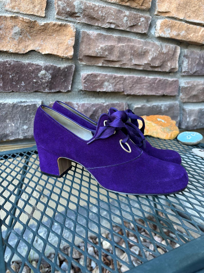 May include: A pair of purple suede shoes with a low heel and lace-up detailing. The shoes have a rounded toe and a small, stacked heel. The laces are a matching purple colour. The shoes are sitting on a metal surface.