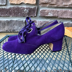 May include: A pair of purple suede heeled shoes with lace-up detailing. The shoes have a rounded toe and a block heel. The laces are a matching purple. The shoes are displayed on a metal surface.
