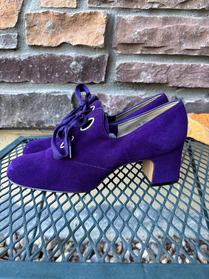 May include: A pair of purple suede heeled shoes with lace-up detailing. The shoes have a rounded toe and a block heel. The laces are a matching purple. The shoes are displayed on a metal surface.
