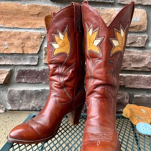 Op de afbeelding: Een paar bruine leren cowboylaarzen met bloemenborduurwerk in geel en crème. De laarzen hebben spitse tenen, hoge hakken en decoratieve stiksels. De laarzen worden tentoongesteld op een metalen rooster.