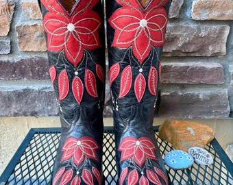 May include: A pair of black cowboy boots with red floral embroidery. The boots feature a pointed toe and a tall shaft, with a decorative flower and leaf design. The boots are made of leather and have a classic Western style.