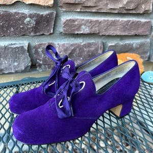 Puede incluir: Un par de zapatos vintage de ante morado con diseño de cordones. Los zapatos tienen un tacón bajo y una puntera redondeada. Los cordones son de color morado a juego y los ojales son plateados. Los zapatos están sobre una superficie metálica.