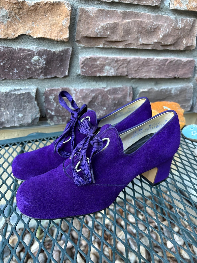May include: A pair of vintage purple suede shoes with a lace-up design. The shoes have a low heel and a rounded toe. The laces are a matching purple colour, and the eyelets are silver. The shoes are on a metal surface.