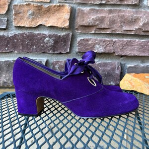 May include: A pair of purple suede shoes with a low heel and ribbon laces. The shoes have a rounded toe and a small heel, approximately 5 cm high. The shoes are sitting on a metal surface.