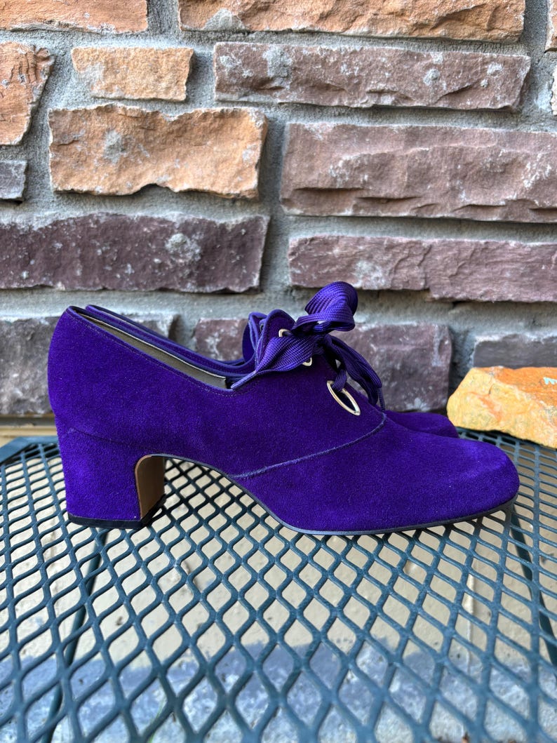 May include: A pair of purple suede shoes with a low heel and ribbon laces. The shoes have a rounded toe and a small heel, approximately 5 cm high. The shoes are sitting on a metal surface.