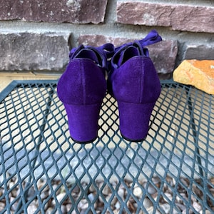 May include: A pair of purple suede shoes with a block heel and ribbon ties. The shoes are set against a brick wall and a metal mesh surface. The heels are approximately 5 cm high.