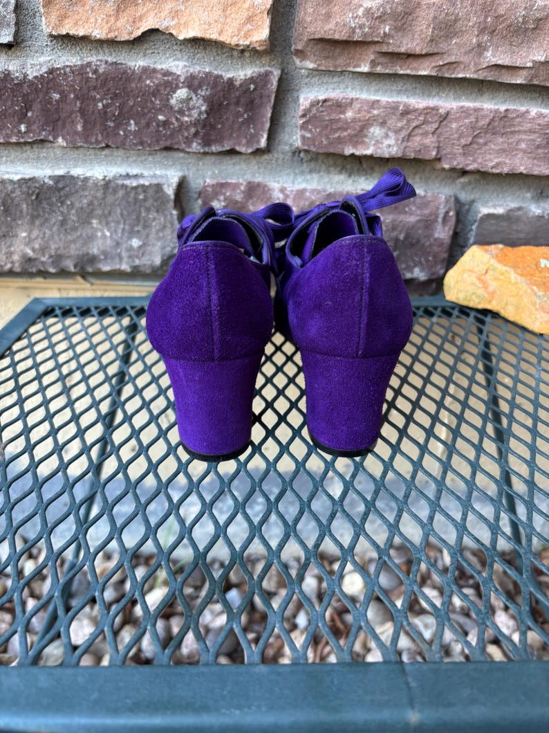 May include: A pair of purple suede shoes with a block heel and ribbon ties. The shoes are set against a brick wall and a metal mesh surface. The heels are approximately 5 cm high.