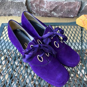 May include: A pair of vibrant purple suede shoes with matching ribbon laces. The shoes feature a rounded toe and gold-toned eyelets. The heels are low. The shoes are displayed on a metal surface.