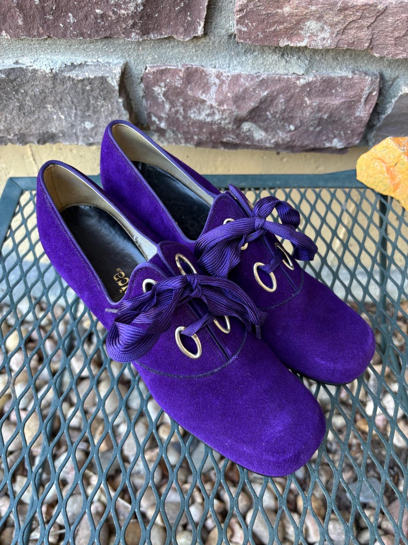 May include: A pair of vibrant purple suede shoes with matching ribbon laces. The shoes feature a rounded toe and gold-toned eyelets. The heels are low. The shoes are displayed on a metal surface.