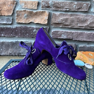 May include: A pair of vintage purple suede shoes with a lace-up design and small heels. The shoes have a rounded toe and are displayed on a metal surface with a brick background.