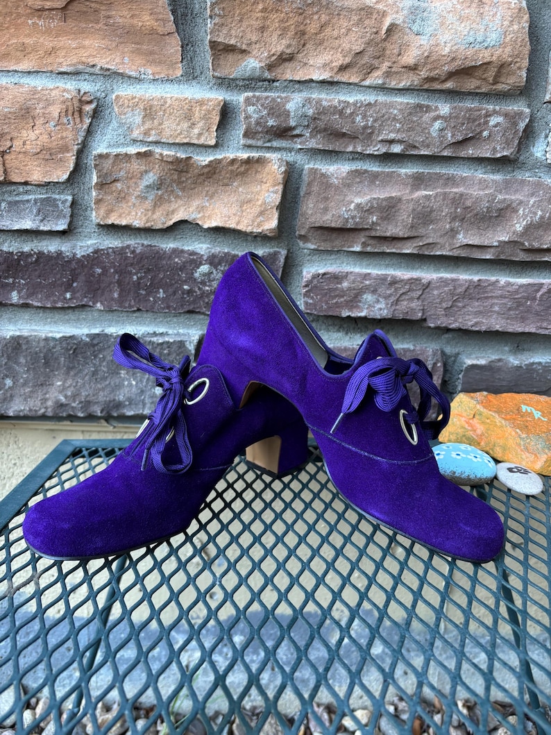 May include: A pair of vintage purple suede shoes with a lace-up design and small heels. The shoes have a rounded toe and are displayed on a metal surface with a brick background.