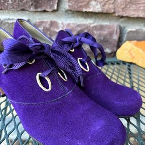 May include: A pair of purple suede shoes with rounded toes and ribbon laces. The shoes have gold eyelets and a low heel. The shoes are displayed on a metal surface with a brick background.