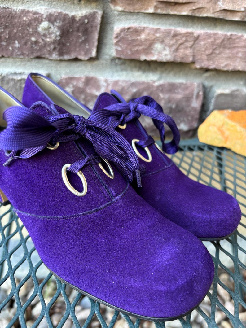 May include: A pair of purple suede shoes with rounded toes and ribbon laces. The shoes have gold eyelets and a low heel. The shoes are displayed on a metal surface with a brick background.