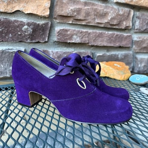 May include: A pair of purple suede shoes with a rounded toe and a small block heel. The shoes have a lace-up design with a satin ribbon and silver eyelets. The shoes are displayed on a metal grate.
