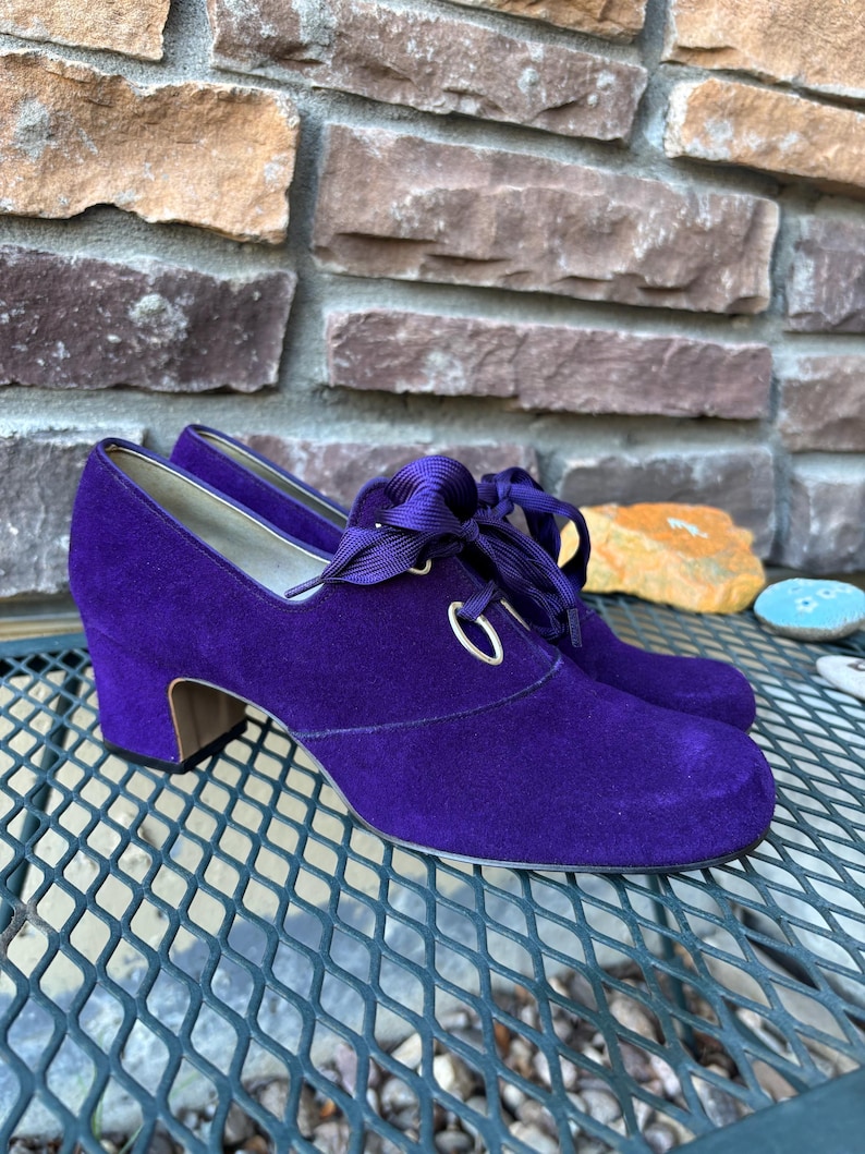 May include: A pair of purple suede shoes with a rounded toe and a small block heel. The shoes have a lace-up design with a satin ribbon and silver eyelets. The shoes are displayed on a metal grate.
