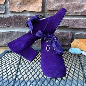 May include: A pair of purple suede ankle boots with a lace-up design. The boots have a pointed toe and a block heel. The laces are a matching purple colour. The boots are displayed on a metal surface with a brick background.
