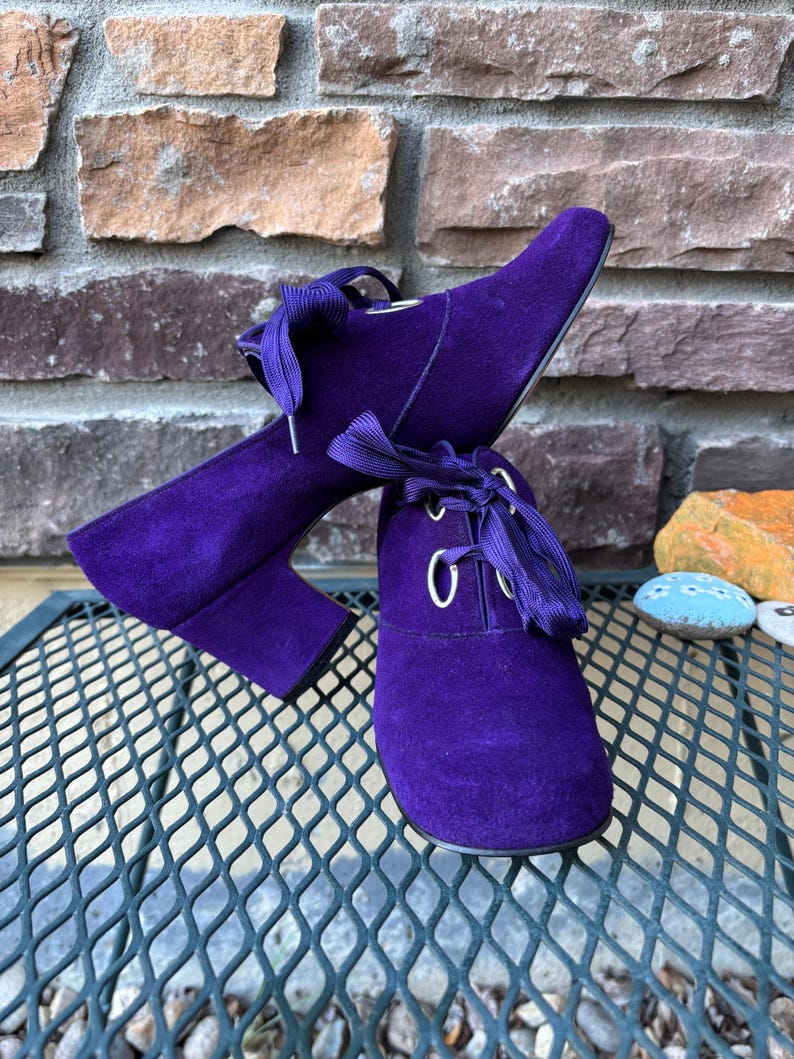 May include: A pair of purple suede ankle boots with a lace-up design. The boots have a pointed toe and a block heel. The laces are a matching purple colour. The boots are displayed on a metal surface with a brick background.