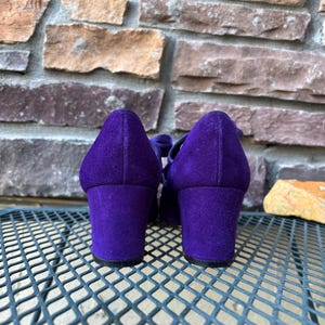 May include: A pair of purple suede shoes with block heels. The shoes have a rounded toe and a strap across the top. The heels are approximately 7.6 cm high. The shoes are set against a textured stone background.
