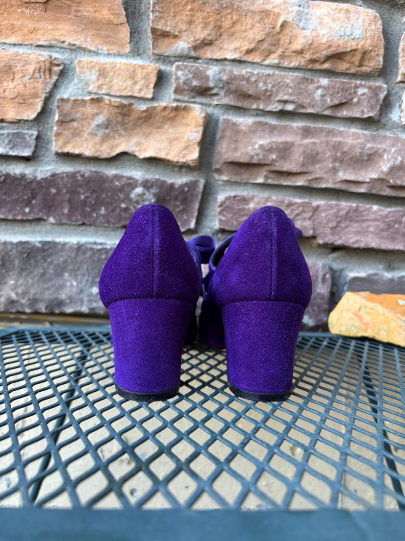 May include: A pair of purple suede shoes with block heels. The shoes have a rounded toe and a strap across the top. The heels are approximately 7.6 cm high. The shoes are set against a textured stone background.