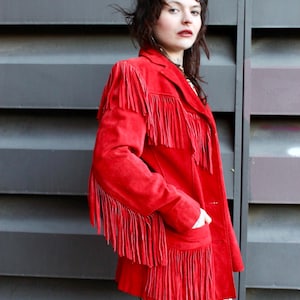 Könnte beinhalten: Eine rote Wildlederjacke mit Fransen. Die Jacke hat einen Knopfverschluss und Taschen. Das Model trägt die Jacke über einem gemusterten Hemd.