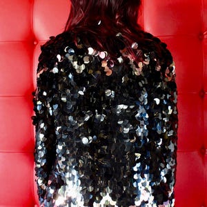 May include: A black sequin jacket with a shiny, reflective surface. The jacket is the focal point, with a red background. The model is wearing leopard print trousers. The jacket is a statement piece.