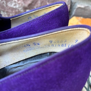 May include: Close-up of a pair of purple suede shoes. The interior of the shoe shows the size, 6 1/2 AA, and other text. The shoes are on a metal surface.