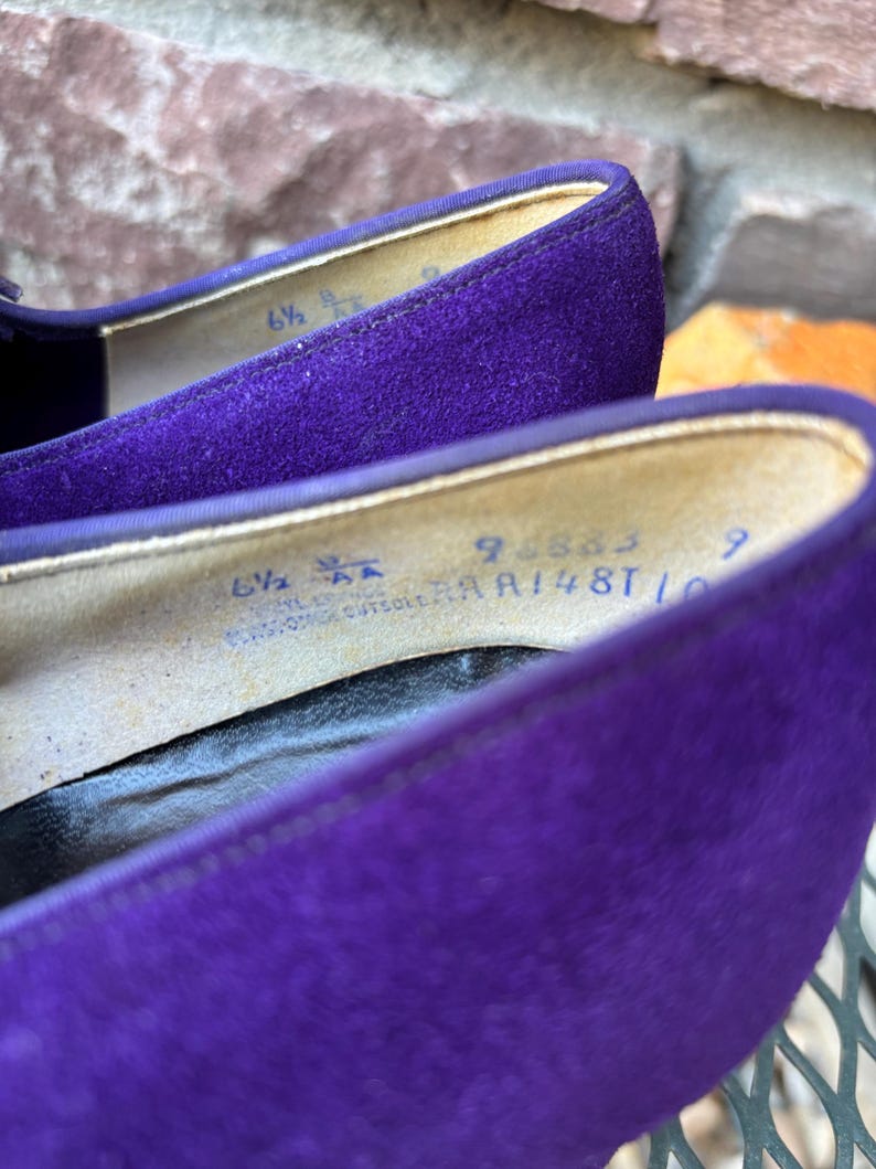 May include: Close-up of a pair of purple suede shoes. The interior of the shoe shows the size, 6 1/2 AA, and other text. The shoes are on a metal surface.