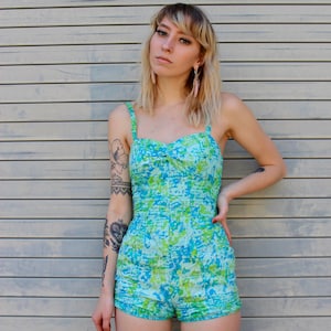 Pode incluir: Uma mulher vestindo um maiô de uma peça com estampa azul e verde com alças finas. O maiô tem um detalhe franzido na cintura e um comprimento curto.