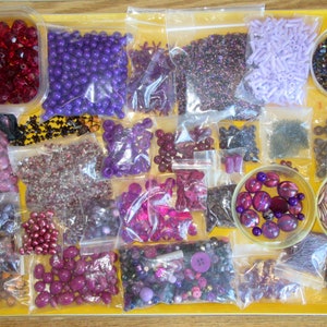 May include: A tray filled with various purple, red, and brown beads and buttons. The beads are in small plastic bags and the buttons are in a small glass bowl. There are also some beads in a larger glass bowl.