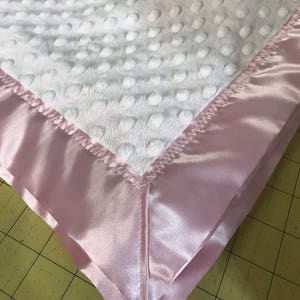 May include: A white baby blanket with a pink satin border. The blanket has a textured, dotted surface.