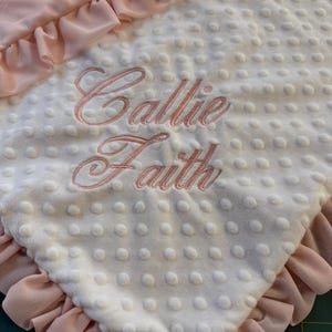 May include: A baby blanket with a white, textured surface and a pink ruffled border. The blanket features the embroidered names "Callie Faith" in a delicate pink script, creating a personalized touch.