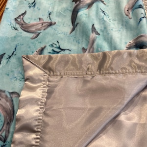 May include: A soft blue and gray blanket with a satin border. The blanket features a repeating pattern of dolphins swimming in the ocean.