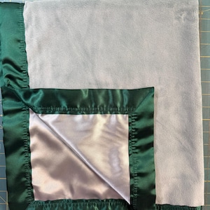 May include: A soft, light gray plush blanket featuring a wide, dark emerald green satin border. The fabric has a smooth, velvety texture on the main surface, while the edges are finished with a shiny, pleated satin trim that frames the blanket.