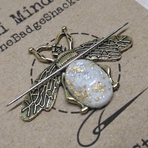 May include: A gold bee pin with white and gold glitter accents. The bee is on a brown card with the text "Mini Mind @ TheBadgeShack" and "stitched".