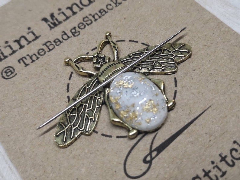 May include: A gold bee pin with white and gold glitter accents. The bee is on a brown card with the text "Mini Mind @ TheBadgeShack" and "stitched".