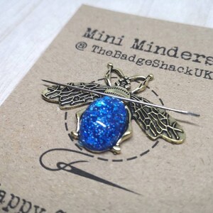 May include: A gold bee-shaped needle minder with a blue glitter centre. The needle minder is on a brown card with the text "Mini Minders @TheBadgeShackUK" printed on it.