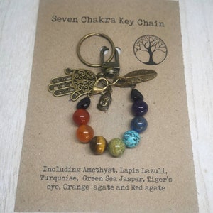 May include: A seven chakra key chain with a hamsa hand charm, a small Buddha charm, and a feather charm. The chain is made of beads in the colors of the seven chakras: purple, indigo, blue, green, yellow, orange, and red. The text on the card reads: "Seven Chakra Key Chain Including Amethyst, Lapis Lazuli, Turquoise, Green Sea Jasper, Tiger's eye, Orange agate and Red agate"